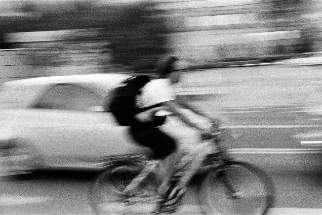 A cyclist passing a car in a blurred black-and-white street scene
